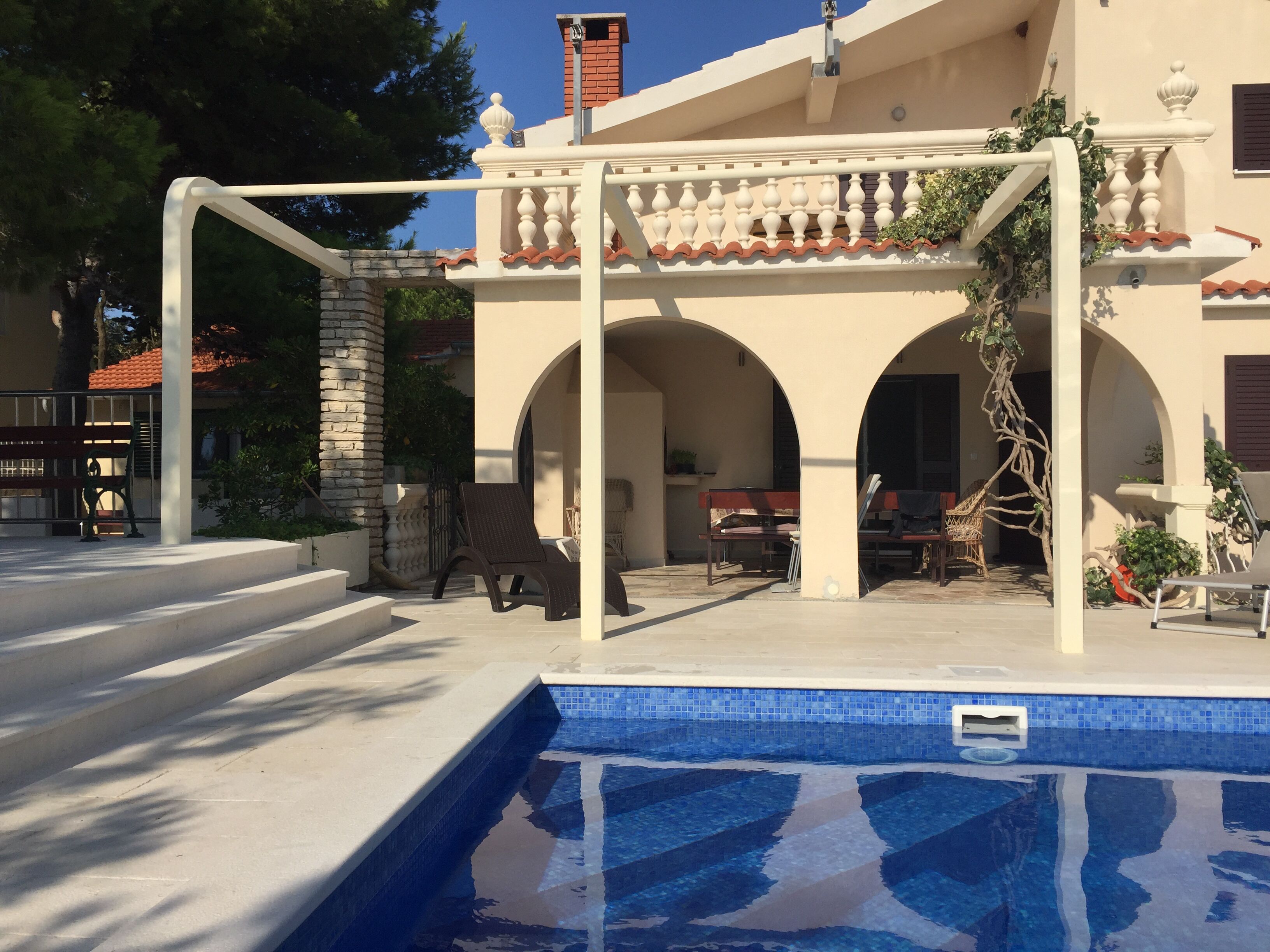 Zona piscina con solarium piastrellato e un pergolato con ringhiere bianche.