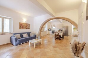 A spacious living area with a sofa, coffee table, and a dining table with chairs. An archway leads to a kitchen area with modern appliances.
