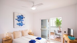 Quarto com cama, ventoinha no teto, pintura de peixes, porta de correr de vidro, escrivaninha com cadeira e uma planta.