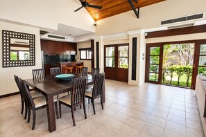 A spacious dining area with a large table and chairs, a kitchen in the background, and a view of a garden through glass doors.