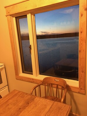 A room with a wooden table and chair, a window with a view of a body of water, and a white appliance in the corner.