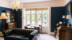 A room with a dark blue wall, a chandelier, a leather sofa, a dining table with chairs, and a window with a view of greenery. - 5 bedroom accommodation in Shap, near Penrith (Shap, near Penrith)