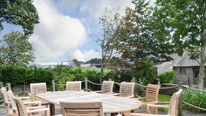 A wooden deck with a round table and chairs, surrounded by greenery and stone buildings. - 5 bedroom accommodation in Shap, near Penrith (Shap, near Penrith)
