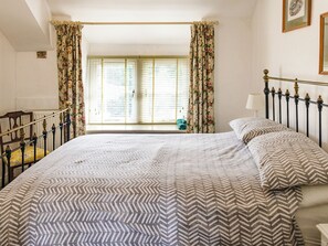 Ein Schlafzimmer mit einem großen Bett, floralen Vorhängen, einem Fenster mit Jalousien, einem Nachttisch mit Lampe und einem gerahmten Bild an der Wand.