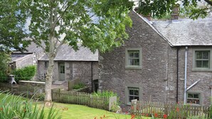 A stone building with multiple windows, a wooden fence, and a grassy lawn with red flowers. - 5 bedroom accommodation in Shap, near Penrith (Shap, near Penrith)