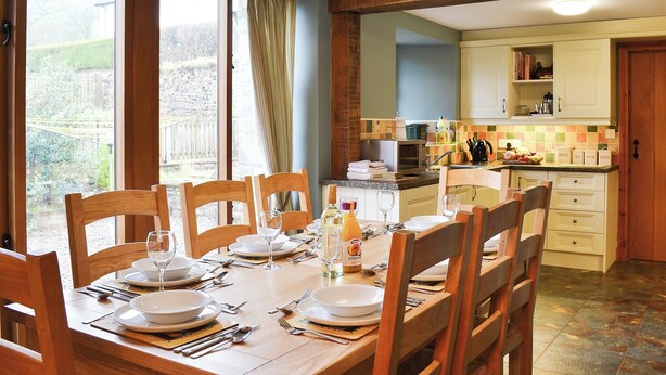 A dining area with a wooden table set for four, a kitchen in the background, and a view of the garden through large windows. - 4 bedroom accommodation in Soar, near Brecon (Soar, near Brecon)