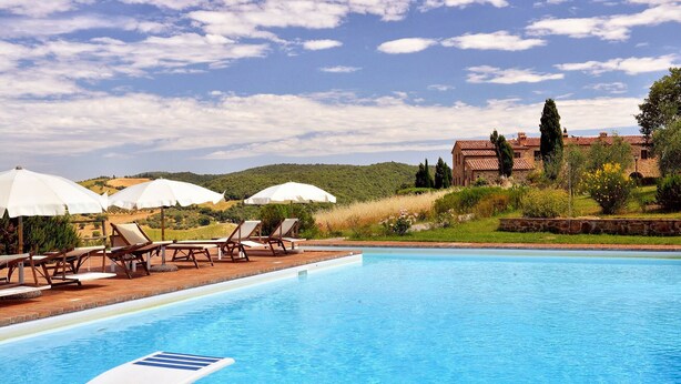 Una piscina con lettini e ombrelloni, immersa nel verde con un edificio sullo sfondo.