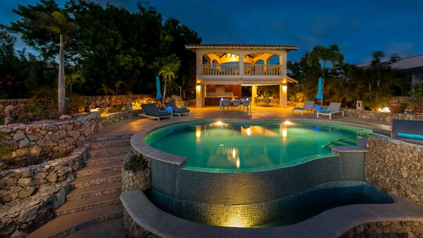 A pool with a tiled edge, a stone wall, and a building with a balcony in the background.