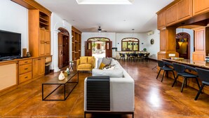 A spacious living area with a large TV, a sofa, and a dining table with chairs. There is a kitchen area with wooden cabinets and a window above the counter.