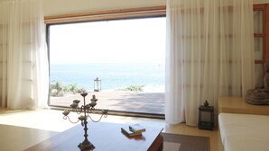 A room with a large window overlooking the sea, a wooden table with a candelabra, and a small bookshelf. - Amazing seafront setting-Infinity pool (Sumartin)