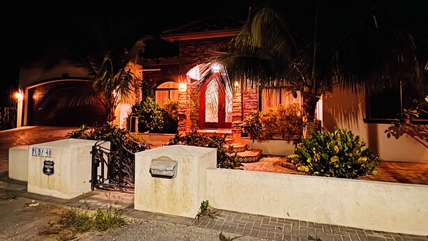 Edifici ben il·luminat amb façana de pedra, una porta vermella i una petita zona de jardí amb plantes i una palmera.