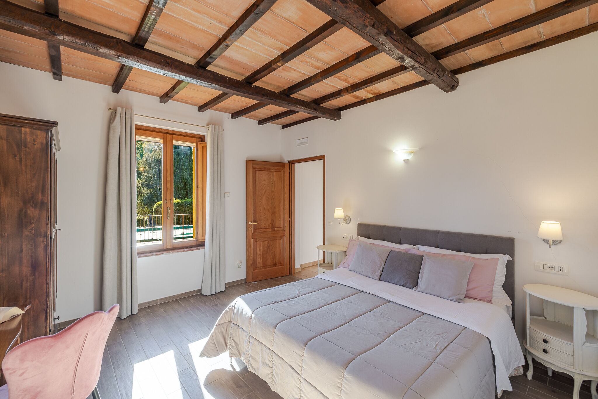 A bedroom with a large bed, wooden ceiling beams, a window with curtains, a bedside table with a lamp, and a chair.