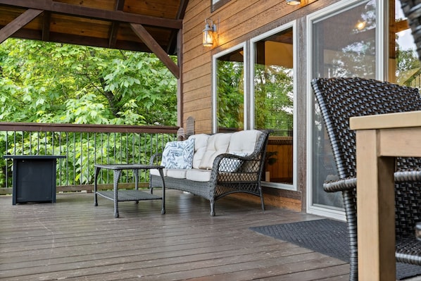 A covered deck with a wicker sofa, a small table, and a trash bin.