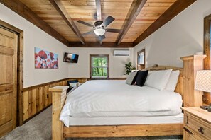 A wooden bedroom with a bed, a ceiling fan, a TV, and a window with a view of greenery. - Mountain Crest Cabin-Relaxing Vacation in Jasper (Jasper)