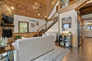 A spacious living room with a high ceiling, wooden beams, a staircase, a fireplace, and a kitchen area with wooden cabinets.