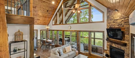 A spacious living area with a high ceiling, wooden beams, a fireplace, and large windows offering a view of the outdoors.