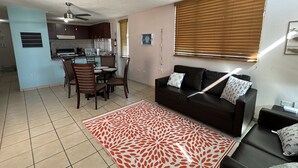 Un salon avec un canapé noir, un coin repas avec une table et des chaises, une cuisine avec des placards et un ventilateur de plafond, et un tapis à motifs sur le sol.