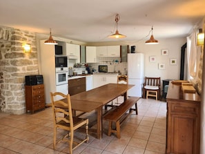 Una cocina con una mesa de madera, sillas e iluminación colgante. En un lado hay una pared de piedra y en el otro, una ventana.