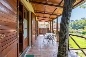 Une terrasse couverte avec une table et des chaises en bois, un tronc d’arbre et une vue sur un paysage verdoyant.
