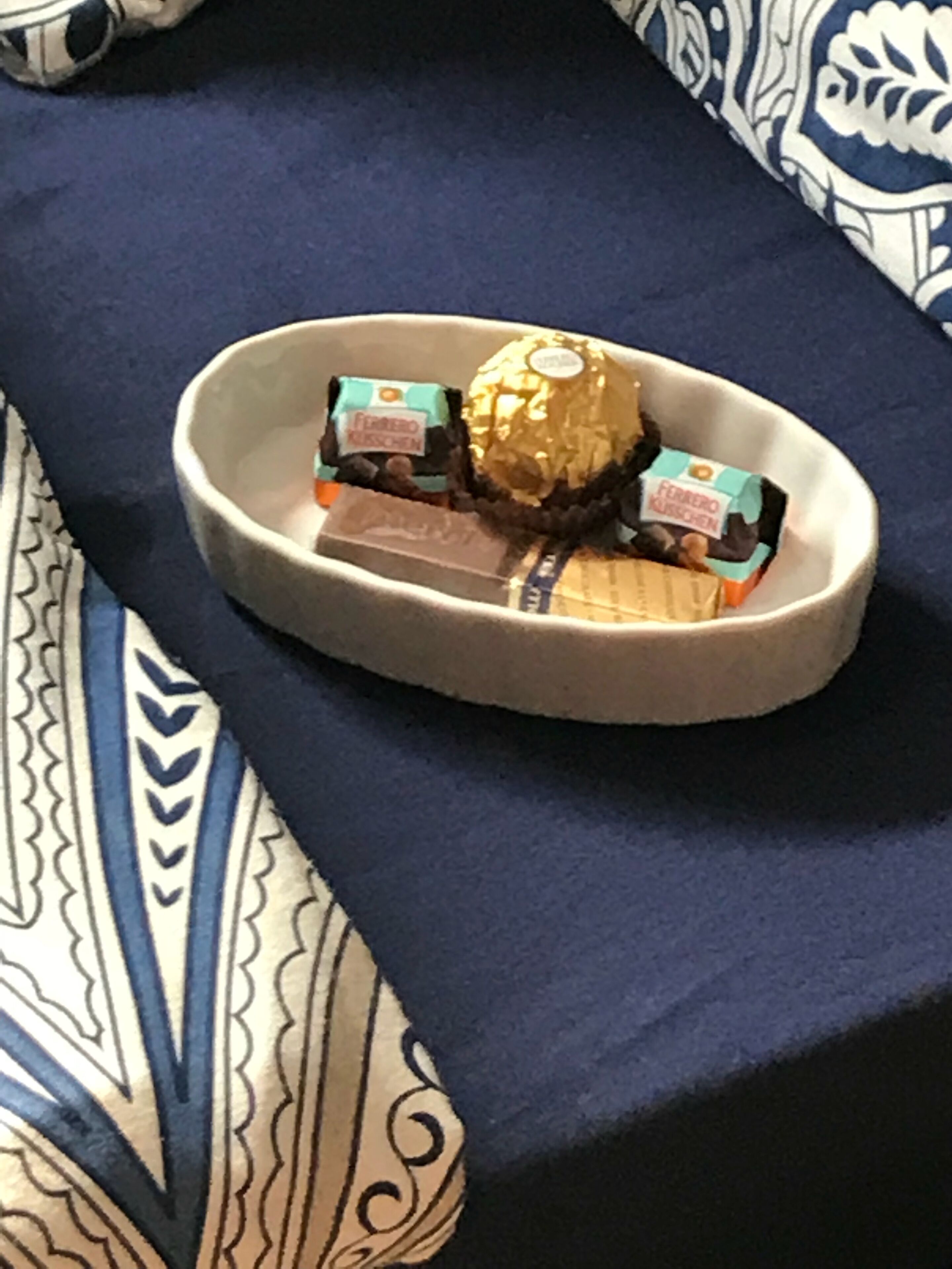 A bed with a blue and white patterned bedspread and a beige tray holding a Ferrero Rocher chocolate and two Ferrero Kauffmann chocolates.