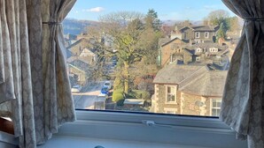 Ein Fenster mit Blick auf eine Stadt, Vorhänge und ein türkisnes Kissen auf einer karierten Sitzunterlage.