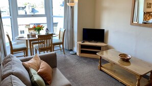 Ein Wohnzimmer mit einer Couch, einem Couchtisch, einer TV-Konsole, einem Esstisch mit Stühlen und einem Fenster mit Vorhängen.