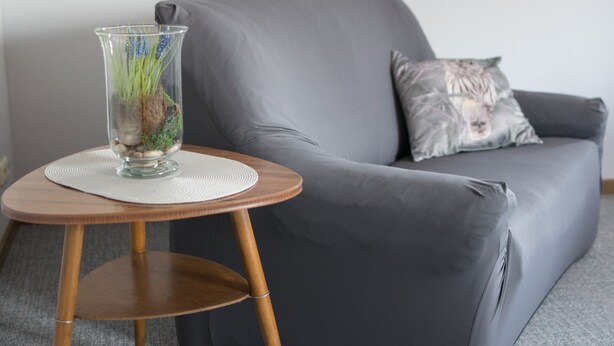 Un sofá gris con un cojín, una mesita de madera con un florero de vidrio y un mantel individual blanco.