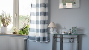 Una habitación con un radiador, una ventana con cortina a rayas, una mesa de vidrio con una lámpara y un cuadro en la pared.
