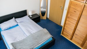 Una cama bien hecha con almohadas y una colcha, una mesita de noche con una lámpara, un armario de madera y un suelo cubierto de moqueta azul.