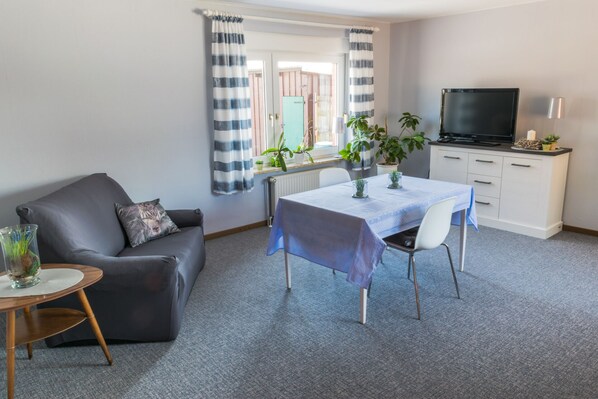 Un salon moderne avec un canapé gris, une table à manger recouverte d’une nappe bleue et un téléviseur posé sur un meuble blanc.