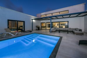 A modern outdoor pool area with a covered seating area and a view of the interior. - Design Villa Maria at Aruba (Noord)