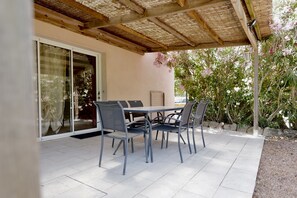 Un patio exterior con mesa y sillas, una pérgola en la parte superior y vista a un jardín con plantas en flor.