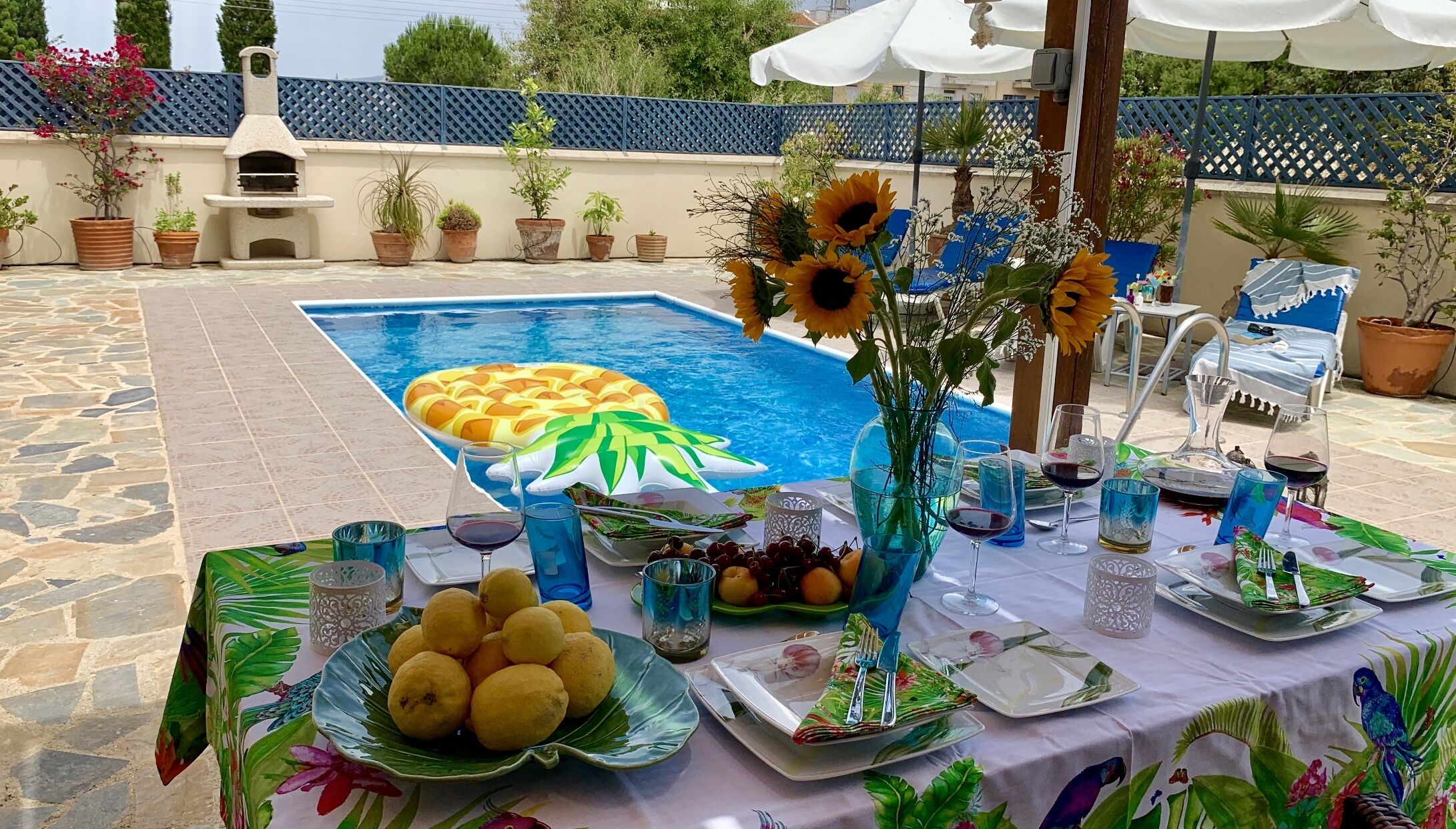 A poolside dining setup with a table set for two, featuring a fruit platter, a vase with sunflowers, and a pineapple float in the pool.