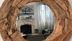 Uno specchio rotondo con una cornice in legno rustico, che riflette un soggiorno con camino, un divano e una finestra con tende.
