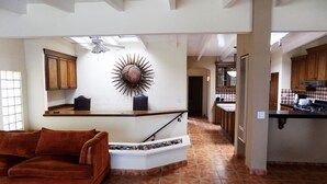 Un'ampia zona giorno con un divano in pelle marrone, un tavolino da caffè bianco e un elemento decorativo a forma di sole sulla parete. Si intravede sullo sfondo una cucina con mobili in legno e un piano di lavoro nero.