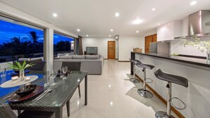 A modern kitchen with a dining area, a living room with a sofa, and a balcony with a view of palm trees.