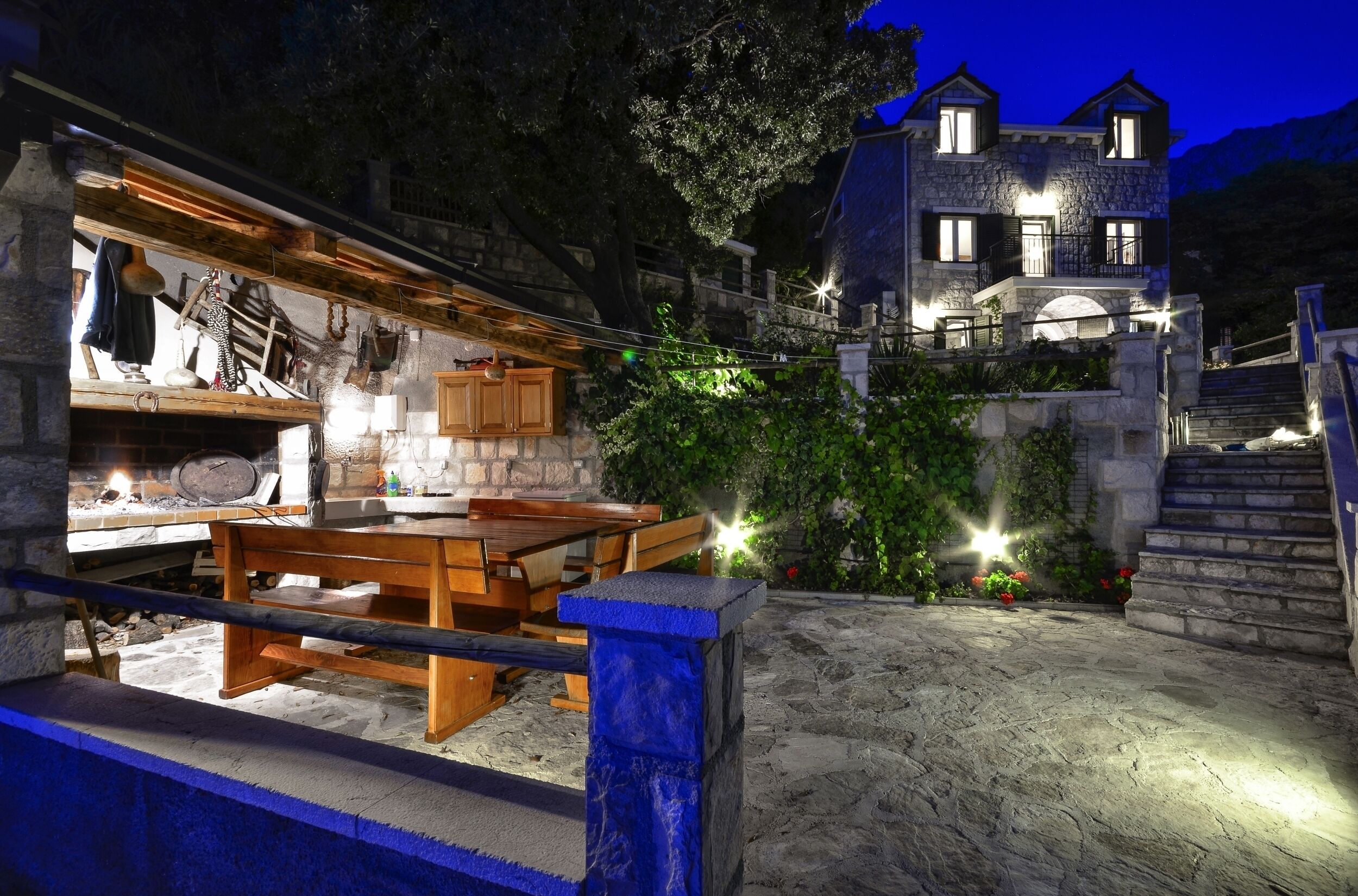 A stone building with a wooden table and benches in the foreground, and a staircase leading to a second-story balcony.