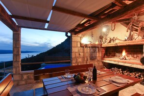 Un espace extérieur aménagé pour les repas, avec un mur en pierre, des bancs en bois, une table dressée pour deux personnes et une vue sur la mer.