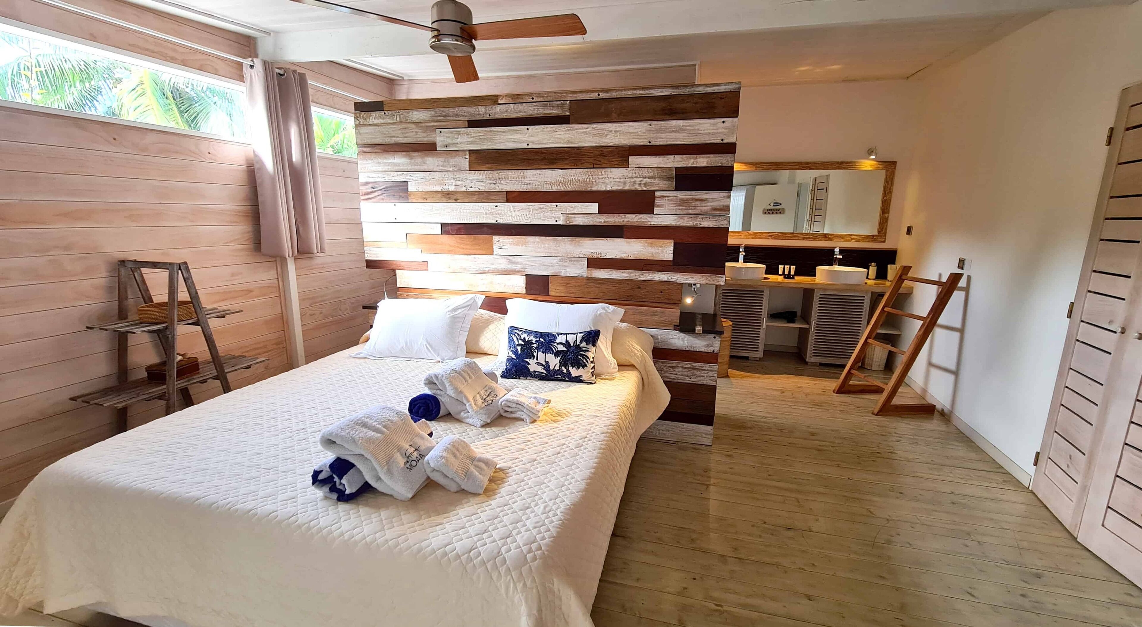 Ein Schlafzimmer mit Holz-Kopfteil, einem Bett mit weißer Bettwäsche, Holzboden, einem Badezimmer mit Spiegel und Waschbecken sowie einem Fenster mit Vorhängen.