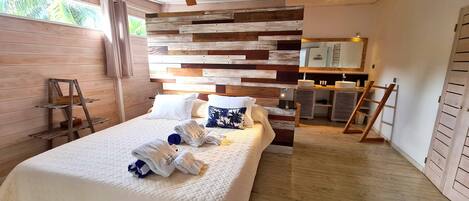 Ein Schlafzimmer mit Holz-Kopfteil, einem Bett mit weißer Bettwäsche, Holzboden, einem Badezimmer mit Spiegel und Waschbecken sowie einem Fenster mit Vorhängen.