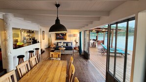 Een ruime binnenruimte met een houten eethoek, een bar met krukken, een bank en een groot raam met uitzicht op een wateroppervlak.