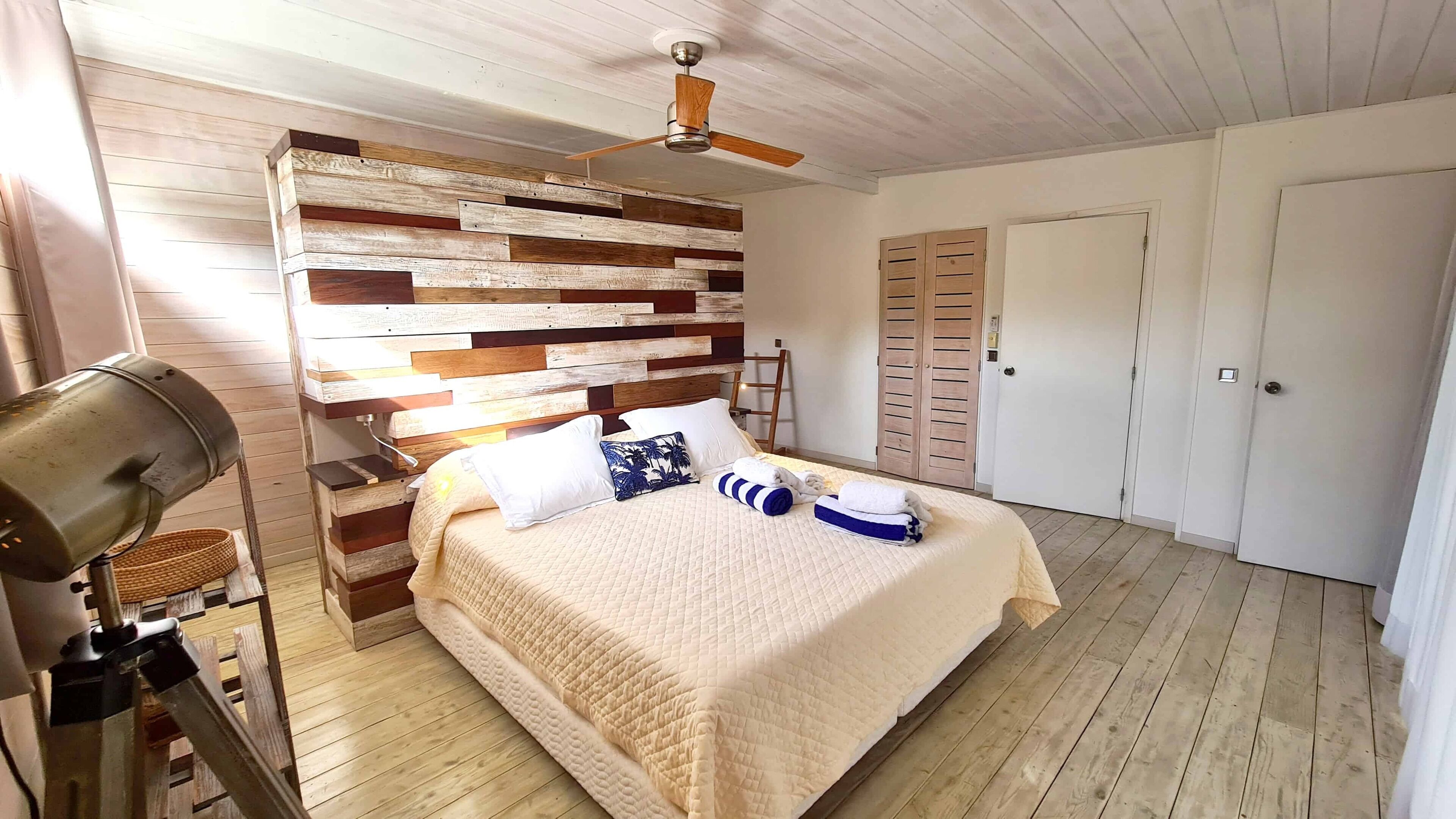 A bedroom with a wooden headboard, a bed with a quilted bedspread, pillows, a ceiling fan, and a wardrobe.