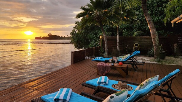 Een houten terras met lounge stoelen en een tafel, met uitzicht op een kalme zee en een zonsondergang.