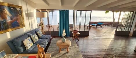 Een ruime woonkamer met een grote bank, een bijzettafeltje en uitzicht op de oceaan door grote ramen.