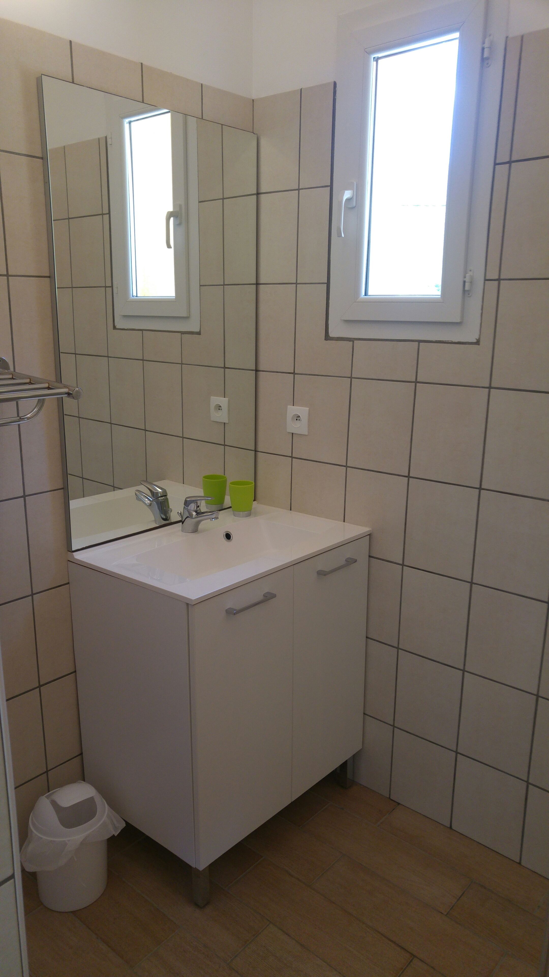 Un baño con un lavabo blanco, un espejo y dos tazas verdes sobre el mostrador. Hay una ventana sobre el lavabo y un cubo de basura en la esquina.