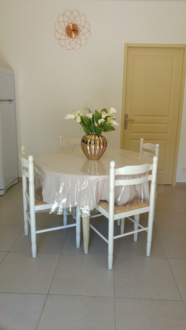 Una mesa redonda para comer con mantel blanco, rodeada por cuatro sillas blancas. Un jarrón decorativo con flores blancas está sobre la mesa.