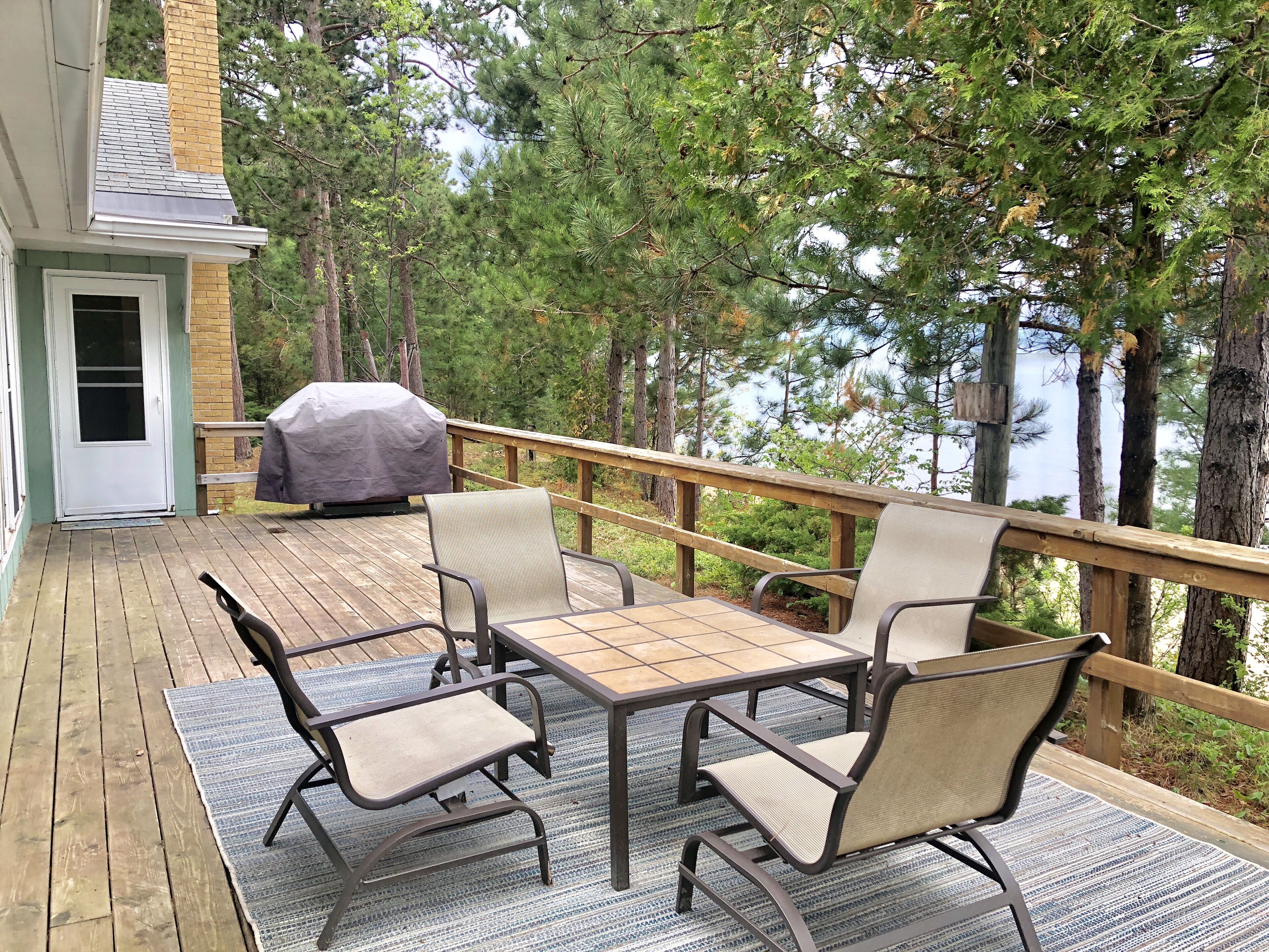 A deck with outdoor furniture, a barbecue grill, and a view of trees and a building.