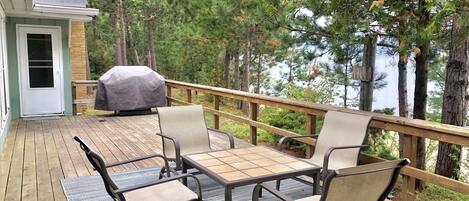 A deck with outdoor furniture, a barbecue grill, and a view of trees and a building.