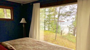 A bedroom with a large bed, a window with a view of trees and a body of water, and a lamp on the left.
