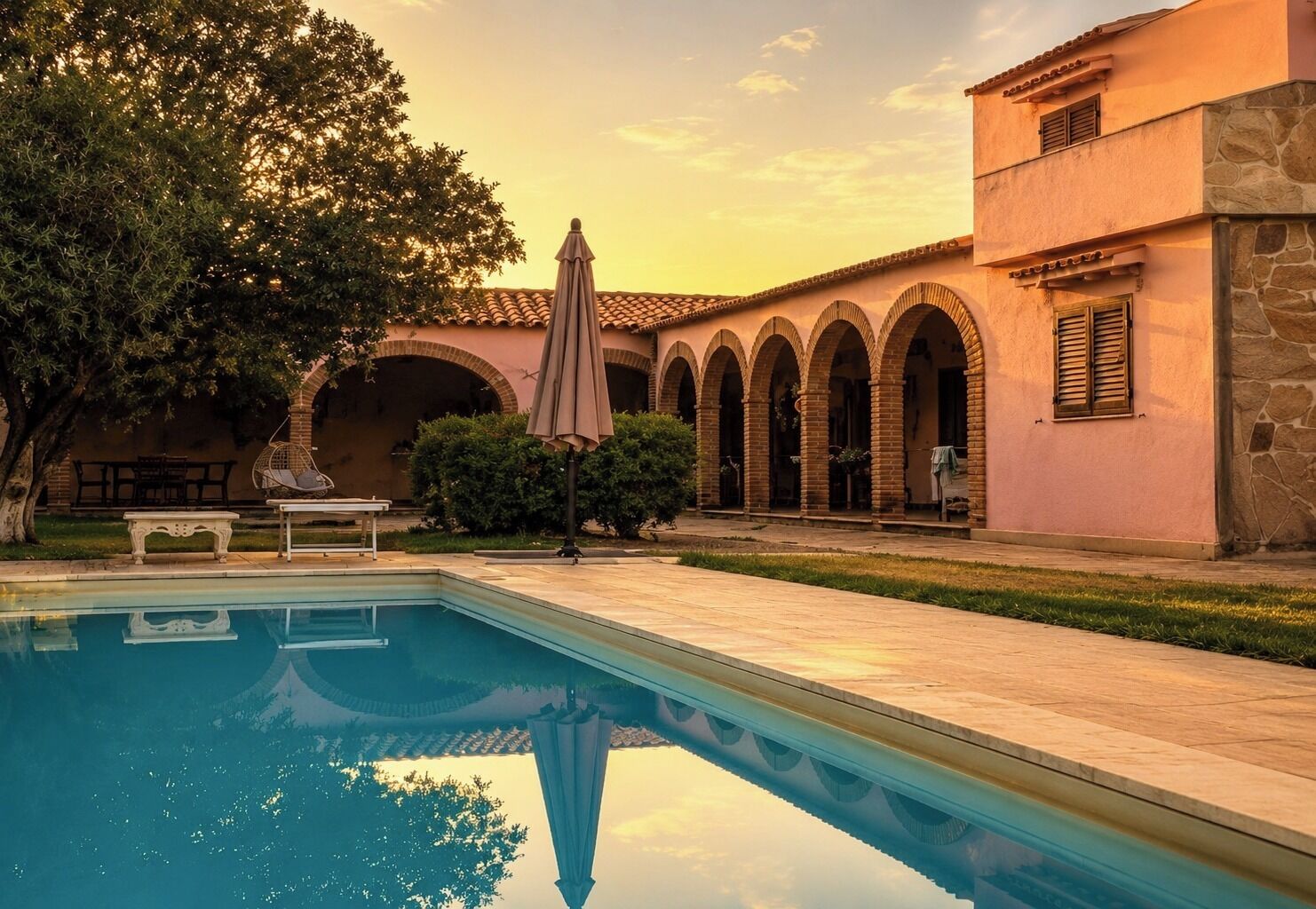 A pool with a clear reflection, surrounded by a building with arched walkways and a patio area.